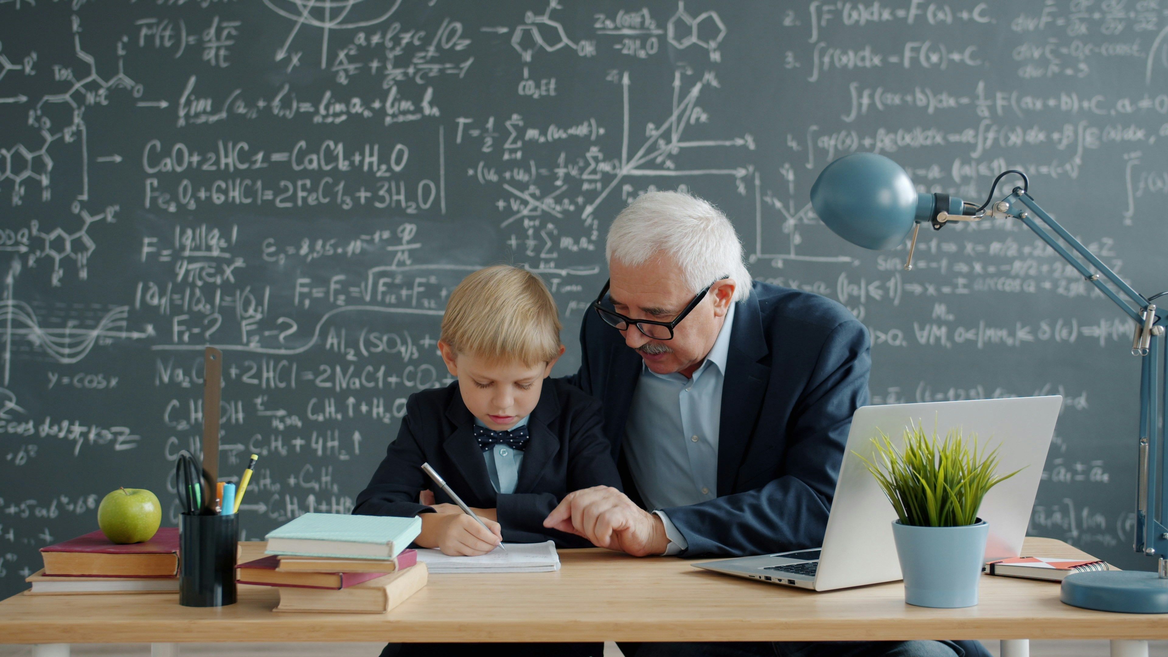Well-dressed older man helping child with homework in front of a chalkboard with equations on it.