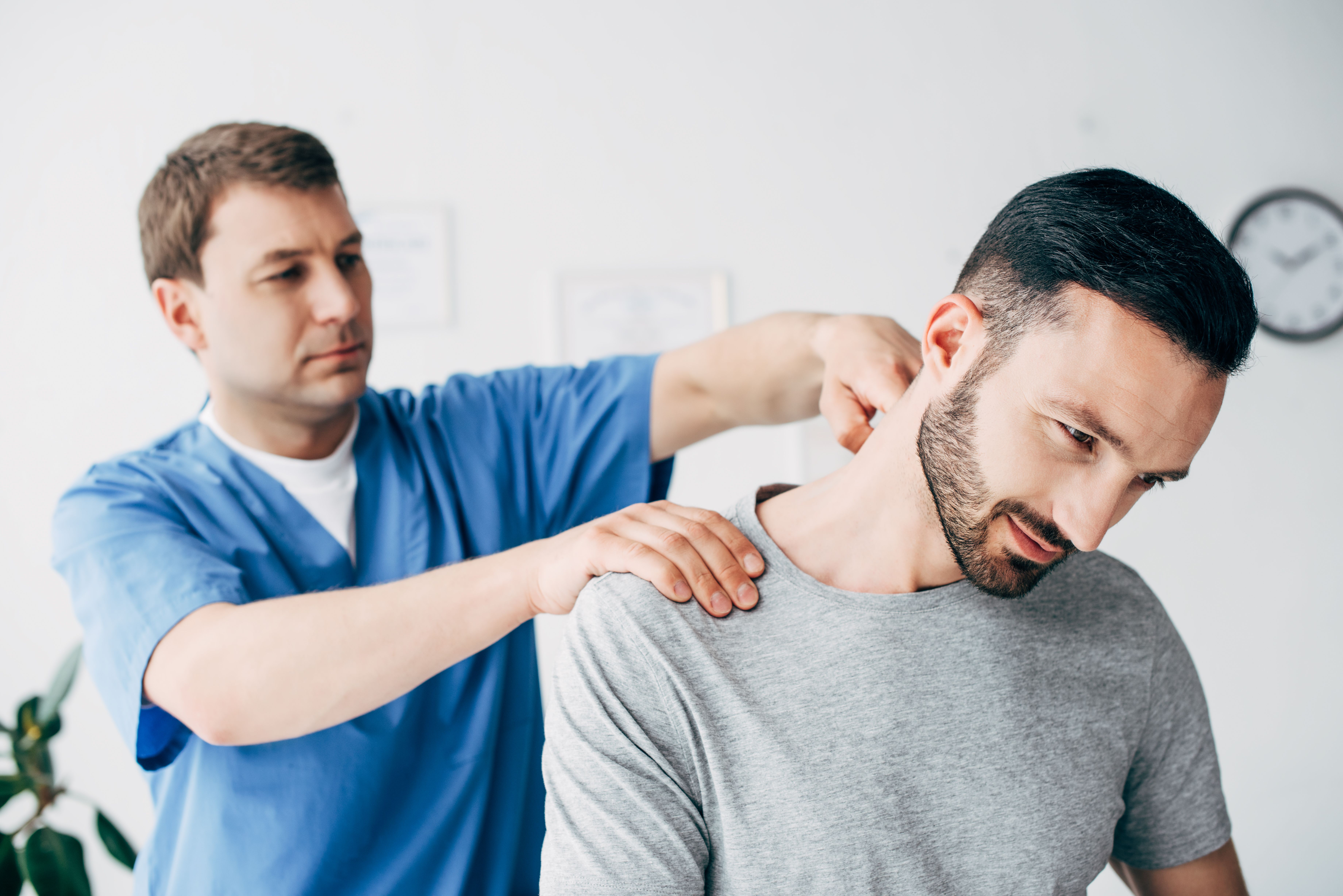 A male healthcare professional is examining a male patient's shoulder and neck area in a clinical setting, attempting to accurately diagnose the cause of the patient's pain.